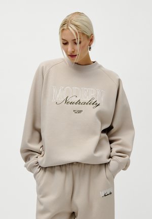 Kvinne med blont hår som har på seg en beige genser og joggebukser med teksten "Moderne Nøytralitet", med hendene i lommer og ser ned.