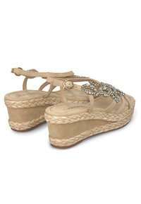 Sandales compensées beiges avec une semelle intermédiaire en jute tressé. Présente des pierres précieuses factices décoratives et des brides de cheville réglables. Texture douce ressemblant à du daim.