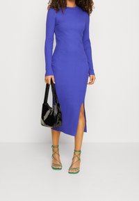 Lång, ribbad, lila klänning med en sidoklyfta, kombinerad med gröna sandaler med remmar och en handväska i svart lack. Har långa ärmar och en rundad halsringning.