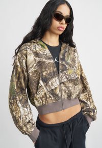 Kvinde iført solbriller, kort camouflage zip-up hoodie, sort Jordan T-shirt og sorte bukser med hænderne i lommerne, poserer mod en ensfarvet baggrund.