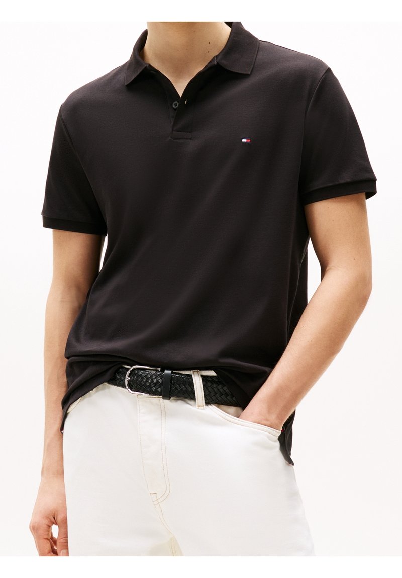 Homme portant un polo noir à manches courtes avec un petit logo sur la poitrine, rentré dans un pantalon blanc avec une ceinture tressée noire.