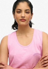 Różowy bezrękawnik wykonany z miękkiego materiału, z zaokrąglonym dekoltem i małym haftowanym logo 'Superdry' po lewej stronie.