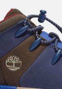 Zapato de senderismo azul y marrón con parte superior texturizada, cordones elásticos y un logo de árbol. Incluye herrajes azules y una lengüeta reforzada.