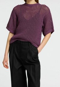 Woman wearing a purple short-sleeve crocheted sweater tucked into high-waisted black pleated trousers against a plain background.