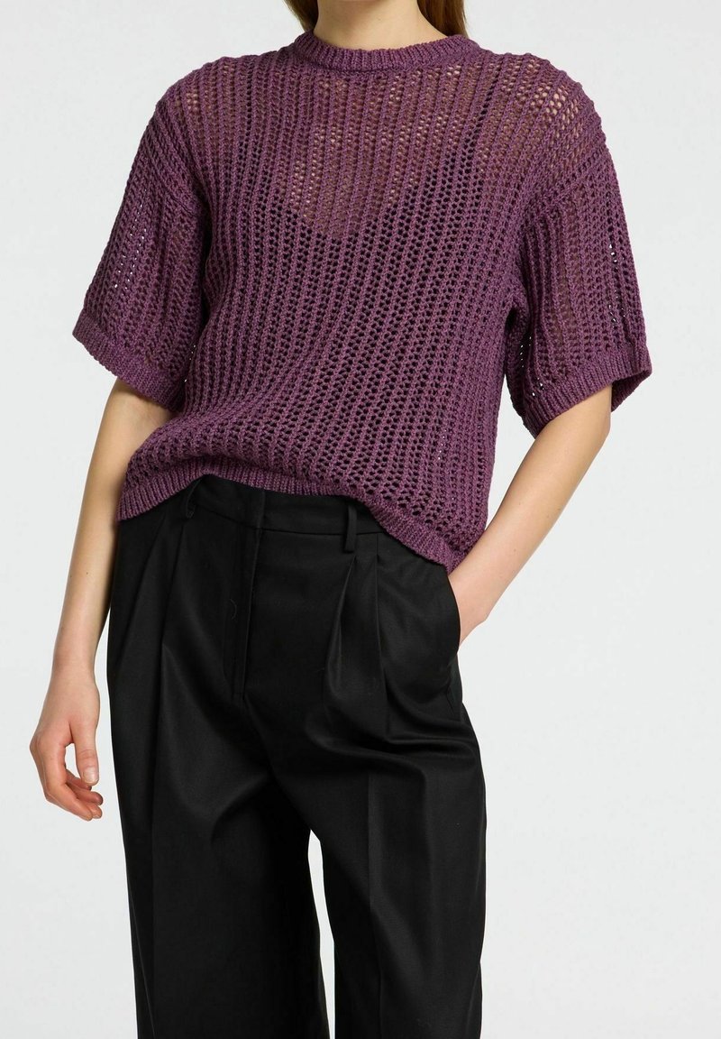 Woman wearing a purple short-sleeve crocheted sweater tucked into high-waisted black pleated trousers against a plain background.
