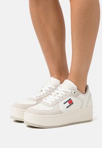 Vita plattformssneakers i mjukt mocka och läder, med en färgglad logotypapplikation på sidan. Platt gummisula med texturerat grepp.