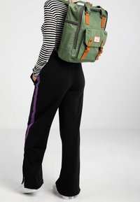 Zelený nylonový batoh s hnědými koženými akcenty; obsahuje přední kapsy, horní uchy a nastavitelné popruhy. Pruhované dlouhé tričko a černé kalhoty.