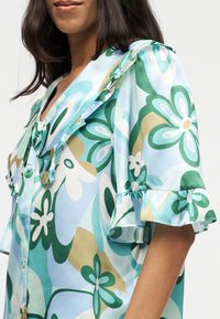 Femme portant un chemisier à manches courtes avec des motifs floraux verts, blancs et beiges, avec un col et des manches à volants sur un fond uni.