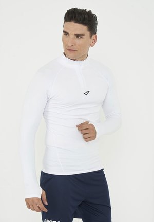 Mann trägt eng anliegendes weißes langärmeliges Sportoberteil mit Daumenlöchern und marineblaue Sportshorts, steht vor einem schlichten weißen Hintergrund.