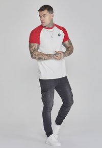 Tricou cu mânecă scurtă, alb și roșu, cu un logo, asociat cu blugi cargo negri și teniși albi. Țesătura pare netedă.