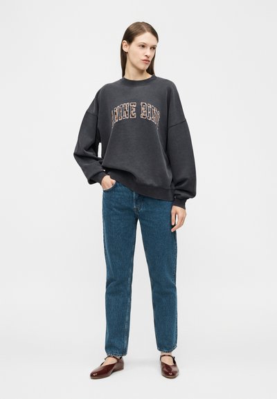 Jauna moteris vilki tamsią, per didelę džemperį su užrašu, mėlynas džinsus ir rudus batelius be kulniukų, stovi viena ranka kišenėje priešais paprastą foną.