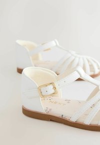 White sandals with a strap design, gold buckle detail, and floral-patterned insole. Matt texture with a low brown sole.