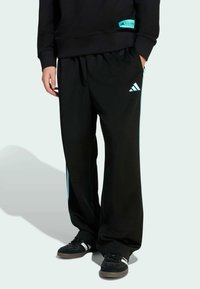 Schwarze Sporthose mit hellblauen Seitenstreifen und Adidas-Logo; verfügt über einen elastischen Bund und eine lockere Passform. Getragen mit schwarzen Schuhen.