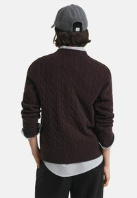 Dunkelbrauner Kabelstrickpullover mit gerippten Bündchen, über einem hellen gestreiften Hemd getragen und mit einer grauen Mütze kombiniert. Einfaches und strukturiertes Design.