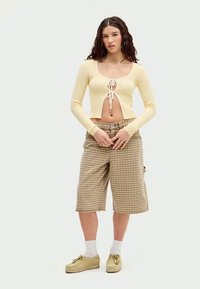Kremljičasti rebrasti crop top s vezanjem, uparen s bež kariranim šortsevima širokih nogavica. Model nosi bež cipele i bijele čarape. Neutralna pozadina.