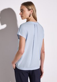 Maglietta leggera blu chiaro a maniche corte con una vestibilità rilassata, caratterizzata da un dettaglio plissettato sul retro e una texture liscia. Abbinata a pantaloni scuri.