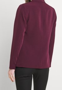 Bordeaux blazer met een gestructureerd ontwerp, voorzien van lange mouwen en subtiele naden, gecombineerd met zwarte aangesloten broek.