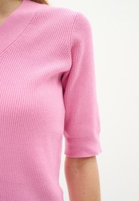 Kvinna i en figurnära, ribbad rosa kortärmad topp med V-ringning mot en enkel, ljus bakgrund.