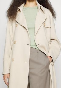 Beżowy dwurzędowy płaszcz z miękkiego materiału, z dużymi guzikami, klapowanym kołnierzem i luźnym krojem, noszony na jasnopistacjowym topie.