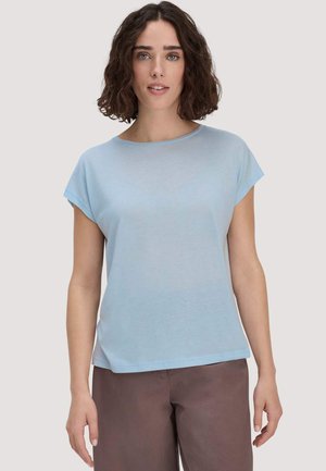 Vrouw met krullend bruin haar tot op de schouders, gekleed in een lichtblauw shirt met korte mouwen en een bruine broek, staand tegen een effen achtergrond.