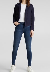 Blazer azul marino de tela texturizada, combinado con una camiseta blanca y vaqueros ajustados de tono azul oscuro, complementado con zapatillas blancas.