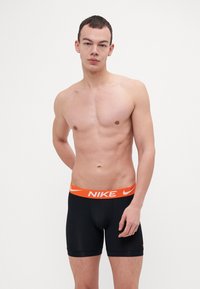 Fekete, testhez simuló boxer alsónadrág narancssárga Nike logóval a derékrészen. Sima szövet, amely szoros illeszkedést biztosít, rövid lábaival és minta nélküli kivitelével.
