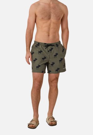 Skjorteløs mand iført grønne badeshorts med sorte elgtrykmønstre og beige sandaler, stående med hænderne i lommerne.