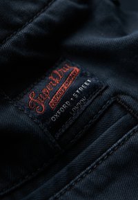 Tela de mezclilla negra con una etiqueta roja y blanca de Superdry que detalla el texto "Oxford Street, Londres." Primer plano con costuras visibles.