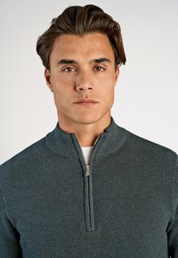Dunkelgrüner Halbzip-Pullover mit strukturiertem Strickdesign und geripptem Kragen, mit einem metallischen Reißverschlussakzent.
