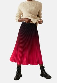 Faltenrock mit Farbverlauf von Schwarz zu lebendigem Rot, kombiniert mit einem beigen, gerippten Pullover und schwarzen Schnürstiefeln. Weiche Stofftextur.