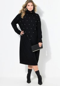 Schwarzes Strickkleid mit Rollkragen, mit Diamantmuster und Perlenakzenten. Kombiniert mit schwarzen Knöchelstiefeln und einer Clutch.