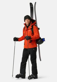 Abbigliamento da sci con giacca impermeabile arancione, pantaloni neri, guanti e scarponi da sci, con gli sci e un casco sulla schiena.