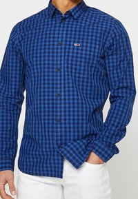 Niebieska koszula w kratę wykonana z bawełny, z kołnierzykiem na guziki i przednią kieszenią, z czarnymi guzikami i małym logo na piersi.
