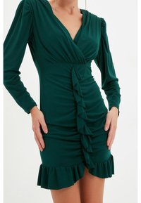 Robe verte à manches longues en tissu doux. Présente un design croisé, un devant froncé avec des volants verticaux et un ourlet froncé.