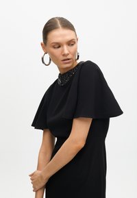 Svart klänning med en pärlbesatt krage, korta svansärmar och en figurnära siluett. Slät textur och elegant design.