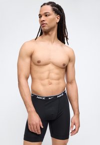 Άνδρας χωρίς πουκάμισο με ντραντλοκς, φοράει μαύρα μπόξερ Nike, στέκεται μπροστά σε απλό ανοιχτό γκρι φόντο, κοιτάζοντας προς το πλάι.