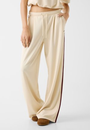 Béžové kalhoty s širokými nohavicemi na stažení v pase a s bočními kapsami, kombinované s hnědými botami, nošené osobou s rukou v kapse.