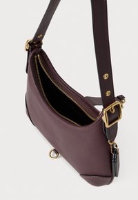 Borsa a spalla in pelle viola scuro con zip e fibbia dorate, tracolla regolabile e piccolo tag nero attaccato da una catena dorata.