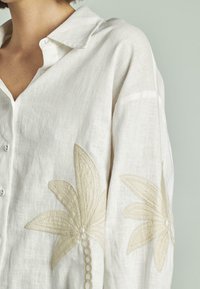 Camicia di lino bianca con colletto, decorata con ricami floreali beige sulle maniche. I bottoni sono visibili lungo la parte anteriore.