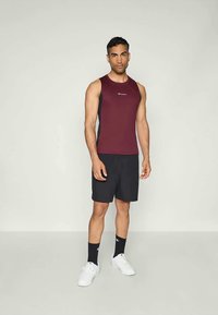 Bordeaux-kleurig mouwloos sportshirt met zwarte zijpanelen, gecombineerd met zwarte shorts. Model draagt witte sneakers en zwarte sokken.