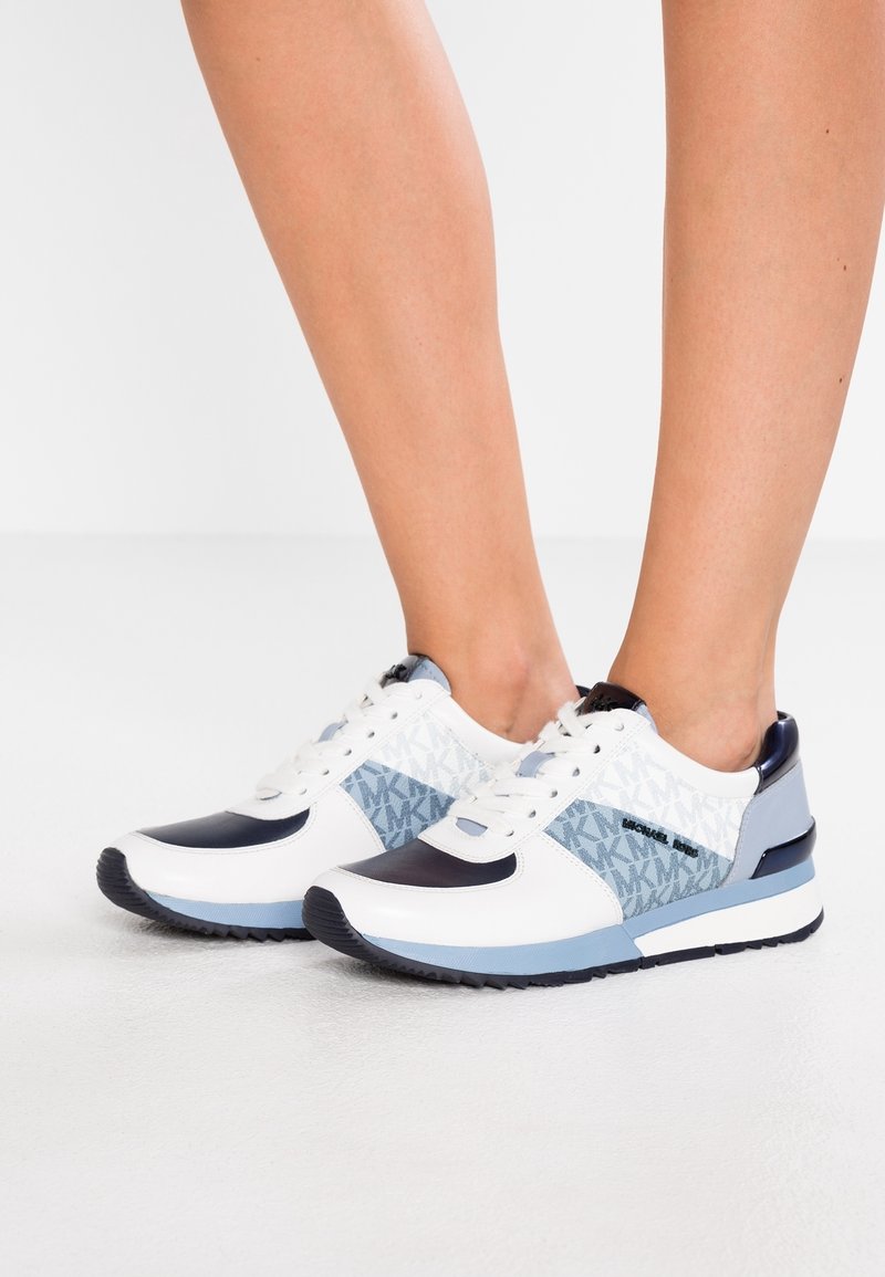Des baskets blanches et bleu marine présentent un panneau bleu texturé avec un motif de logo, des lacets blancs, et un design sportif avec des semelles en caoutchouc.