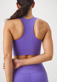 Violetti urheilu crop-top, jossa on urheilullinen taakse avoin malli, sileä rakenne ja tiukka muoto. Yhdistetty vastaaviin korkeavyötäröisiin leggingseihin.