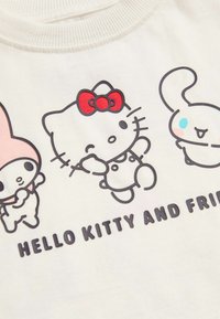 Krémové tričko s potiskem Hello Kitty a jejích přátel, s červenými akcenty a orámovanými detaily. Text zní "HELLO KITTY A PŘÁTELÉ."