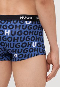 Blauwe boxershorts met herhaaldelijk "HUGO"-tekst in zwart en wit, met een zwarte tailleband waarop de merknaam prominent is weergegeven.