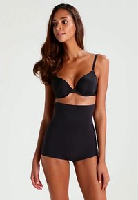 Vrouw met lang bruin haar in een zwarte onderlinge beha en hoog uitgesneden zwarte shapewear shorts, poserend tegen een effen lichte achtergrond.