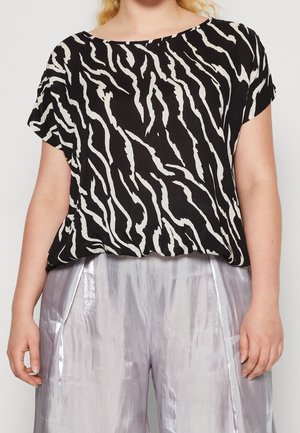 Femme portant un haut à manches courtes imprimé zèbre noir et blanc, associé à un pantalon large argenté brillant, debout devant un fond uni.