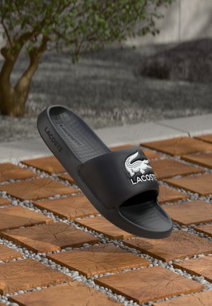 Zwarte rubberen slidesandalen met een textuur in de voetbed en het witte Lacoste-logo. Gladde bandontwerp zonder extra patronen of accenten.