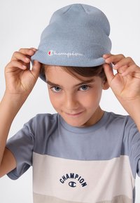 Niño con ojos azules ajustándose un gorro Champion azul claro, vistiendo una camiseta Champion a rayas azul y beige, sonriendo ligeramente contra un fondo blanco.