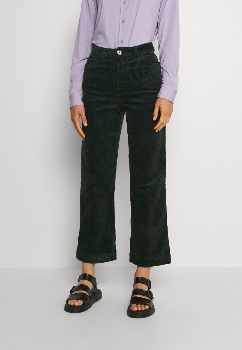 Pantalon large en velours côtelé vert foncé avec une fermeture à boutons à l'avant, assorti à une chemise à manches longues violette claire boutonnée et des sandales noires.