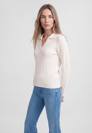 Donna con lunghi capelli biondi che indossa un maglione crema a trecce con colletto e jeans blu, in piedi davanti a uno sfondo chiaro e semplice.